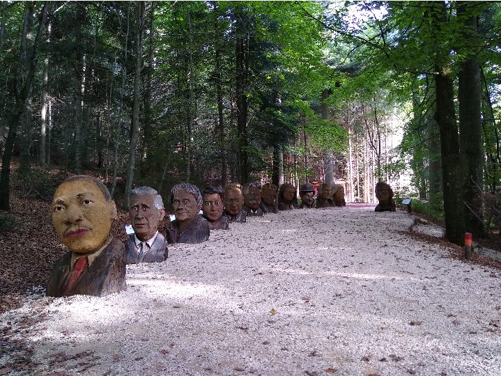 Es ist ein Schotterweg zu sehen. Auf der linken Seite stehen aus Baumstämmen geschnitzte Holzköpfe von berühmten Personen.