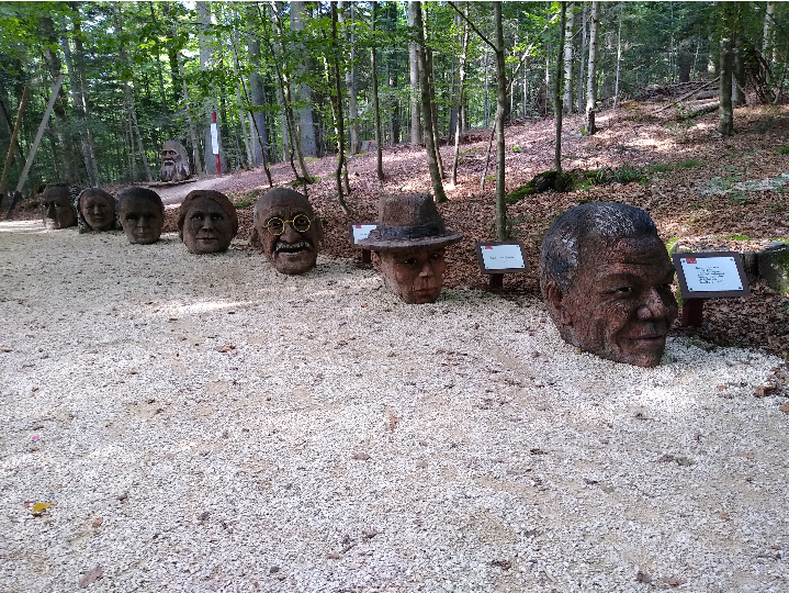 Es ist ein Schotterweg zu sehen. Auf der rechten Seite stehen aus Baumstämmen geschnitzte Holzköpfe von berühmten Personen.