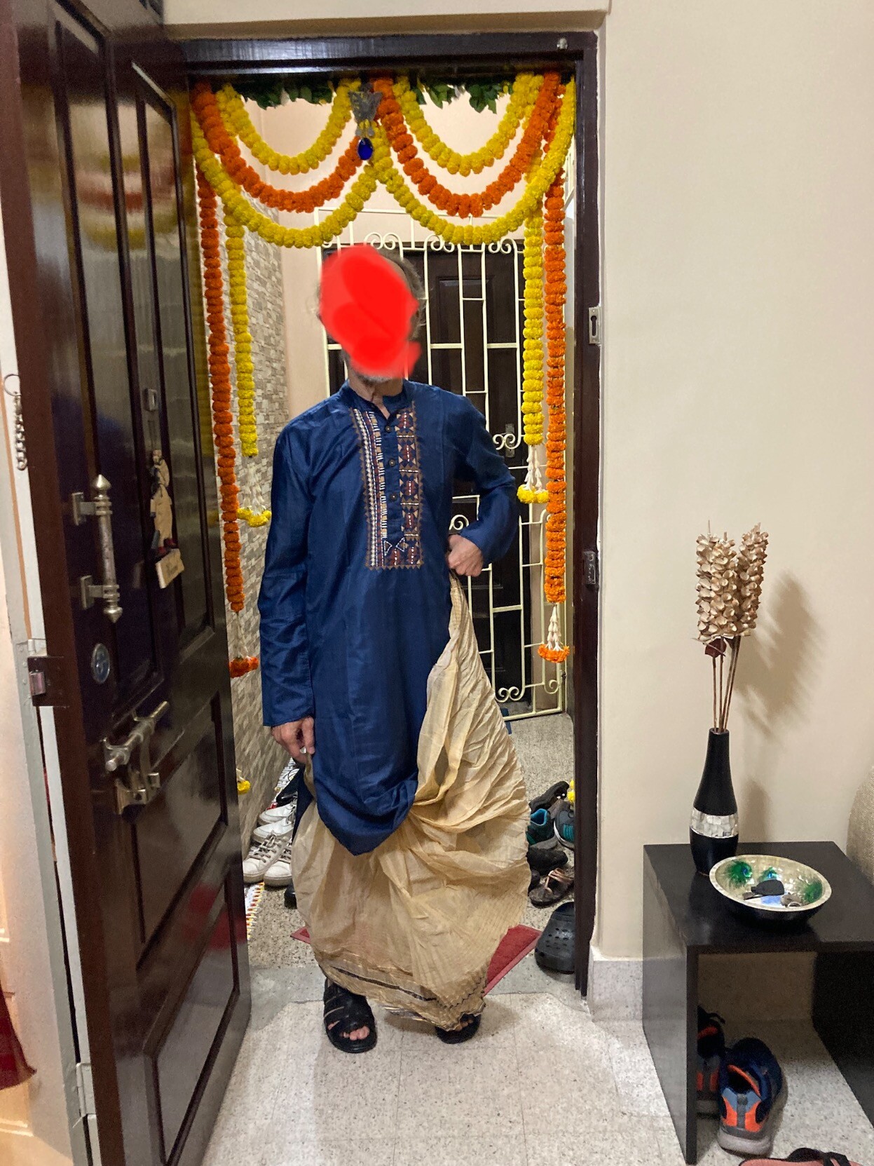 Me standing in a garlanded doorway wearing a dark blue embroidered kuta over a frilly dhoti, holding one end of the latter as one does