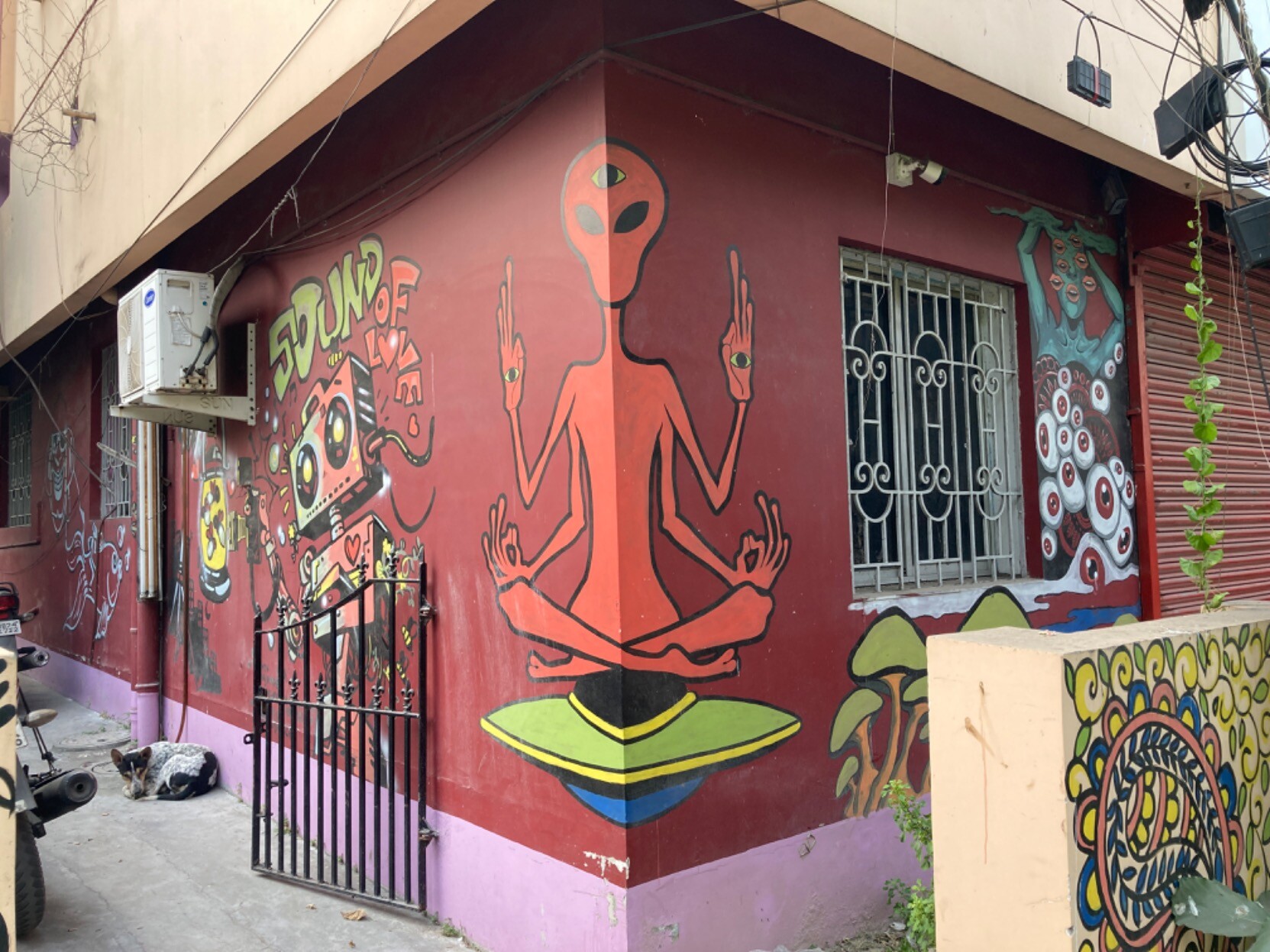 Blood-red building painted with hippiesque designs, e.g. space alien with four arms and eyes in palms sitting in lotus position