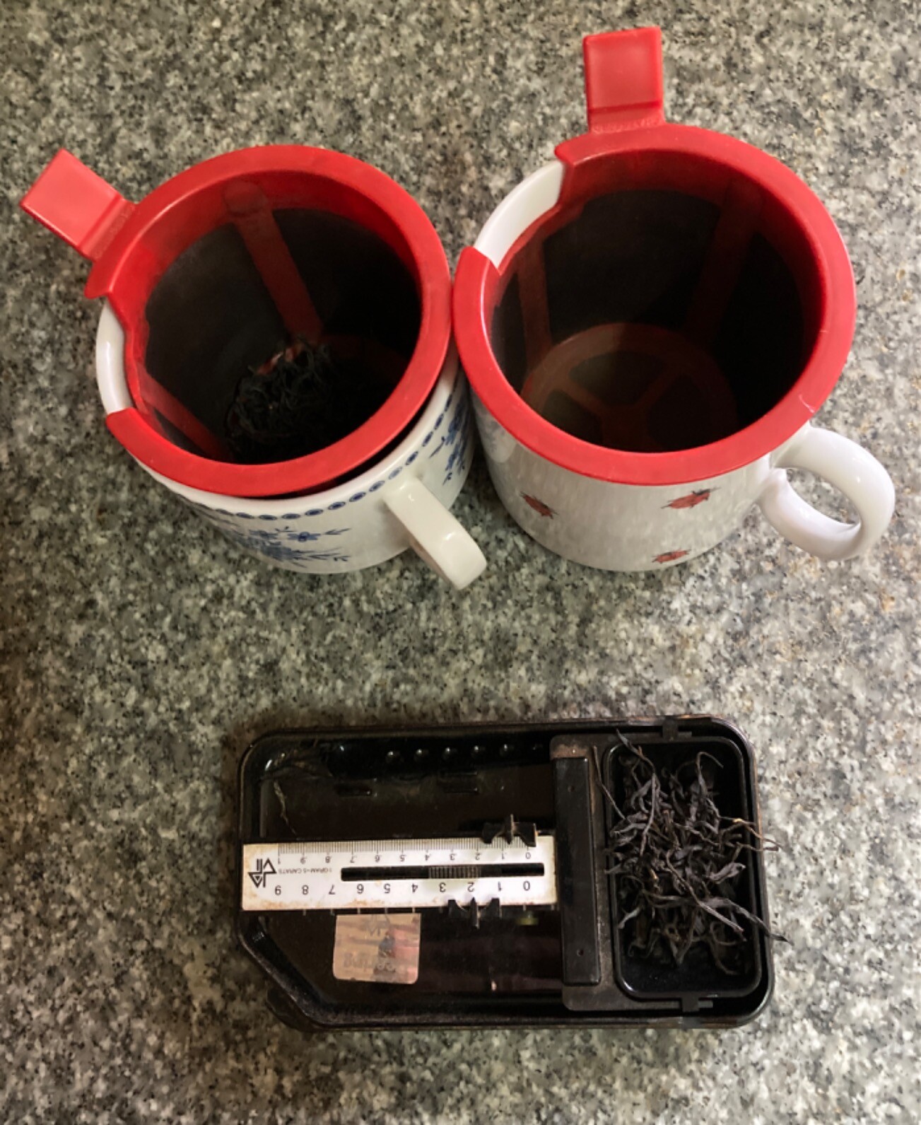 On a granite countertop, two mugs with plastic infuser baskets and a pocket balance scale