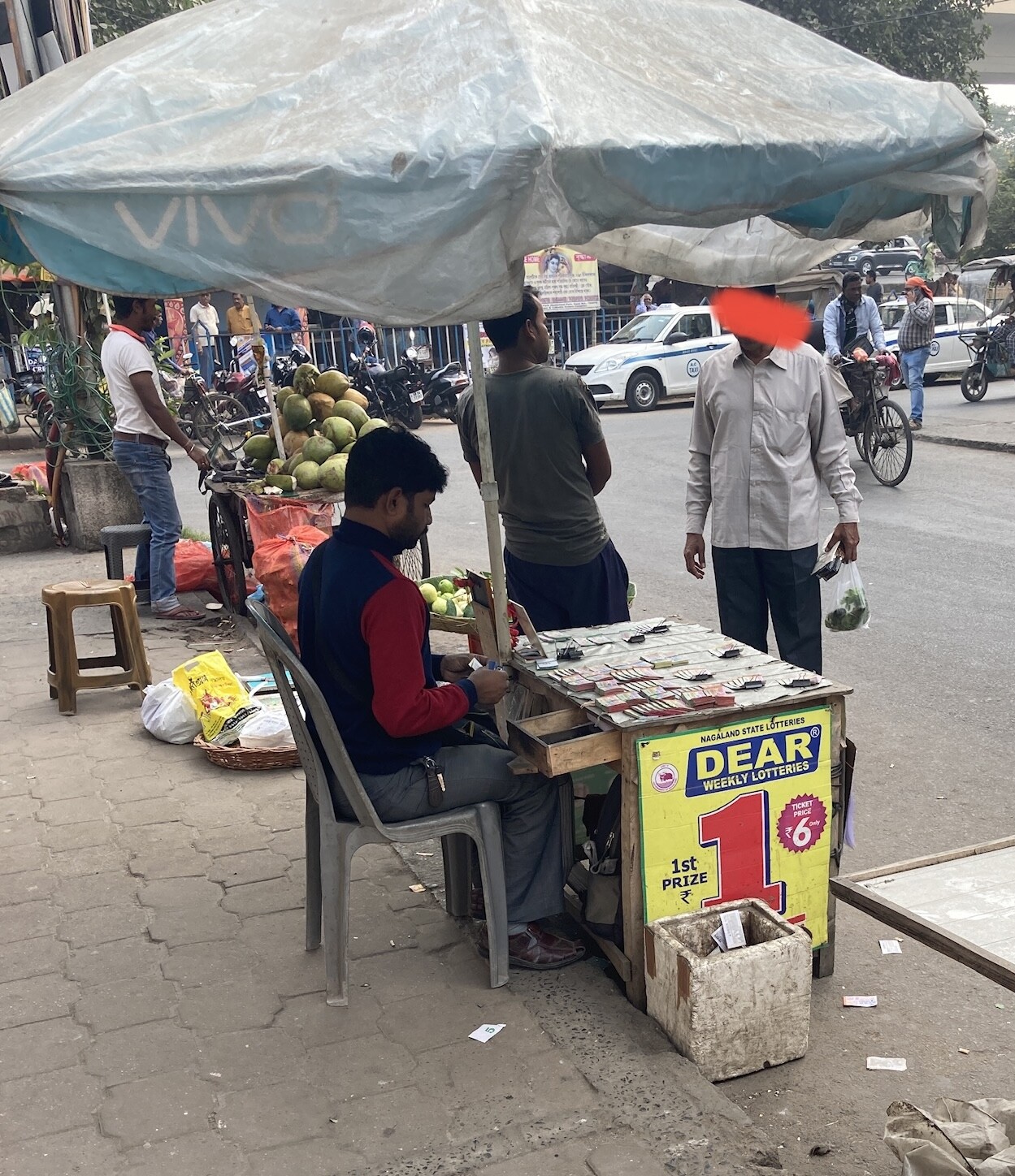 Ticket seller seated at folding table on sidewalk under umbrella, customer in front of him