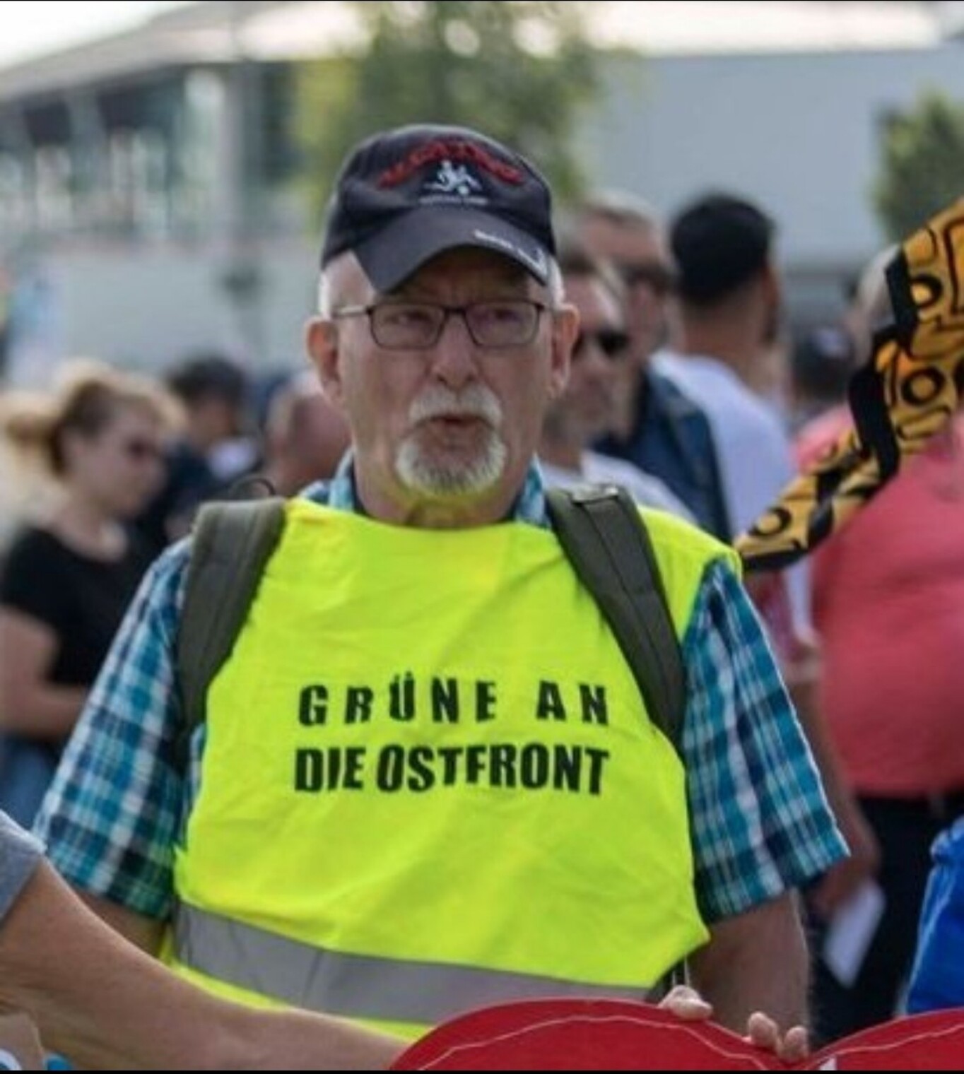 Ein Bild von der Demo in Erdingen heute: ein älterer Herr mit grauem Kinn-Bärtchen und Basecap trägt eine signalgrüne Warnweste über einem karierten Hemd, Aufschrift "Grüne an die Ostfront"