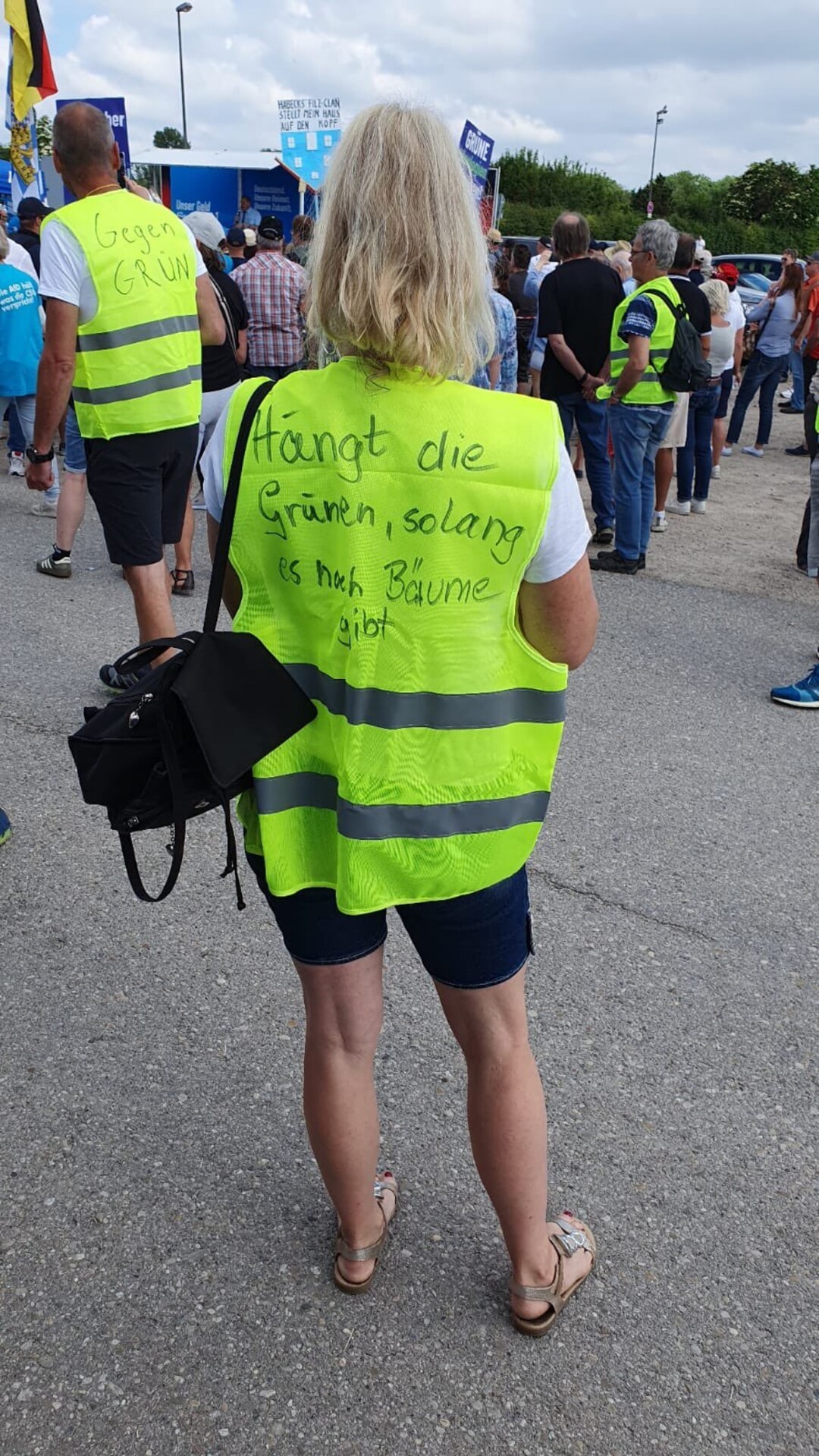 Ein Bild von der Demo in Erdingen heute:<br>Eine Frau mit langen, blonden Haaren in blauen Radlerhosen trägt über ihrer weißen Bluse eine signalgelbe Warnweste. Sie ist von hinten zu sehen, die Audschrift auf dem Rückenteil lautet<br>"Hängt die Grünen, solange es noch Bäume gibt"