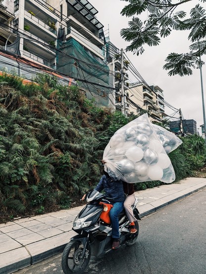 A motorbike on a small road with a large elevated road behind, holding up a bag full of white balloons. Grey sky, of course. Construction and cables on top. Faces of the driver and his companion are hidden behind the balloons 