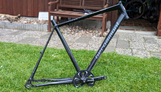Ein schwarzer Fahrradrahmen ohne Gabel und weitere Anbauteile steht auf einer gr+nen Rasenfläche vor einem Geräteschuppen aus Holz.
