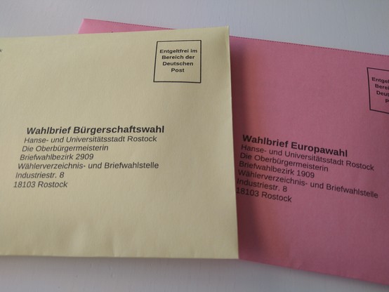 Zwei verschlossene Wahlbriefumschläge, einer in Gelb für die Rostocker Bürgerschaftswahl und einer in Rosa für die Europawahl.