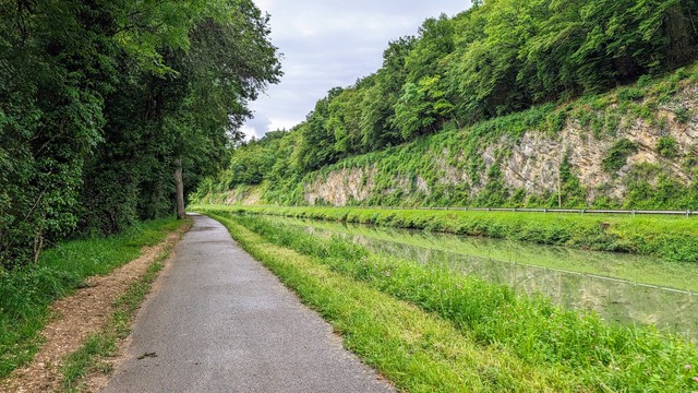 Von links nach rechts: eine Reihe Bäume bildet eine grüne Wand. Dann folgt ein gerader, hinten nach links abbiegender Asphaltweg, dann ein dazu paralleler Kanal und danach eine steile Felswand, auf der Bäume stehen.