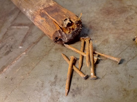 An old wooden handle, covered in rust and grime. A pile of rusty nails is next to the handle, which shows holes and cracked wood where they used to be. The scene rests on a scratched old metal box with grey paint.