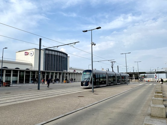 A modern station building with a huge, paved station square and a grey tram running across.