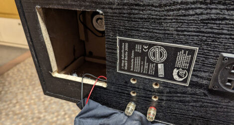 Eine auf der Seite liegende, schwarze Lautsprecherbox mit einem Loch in der Rückseite und 6 Bohrungen für die Anschlussterminals. Zwei der 6 Bohrungen sind mit Lautsprecherkabelbuchsen bestückt.
