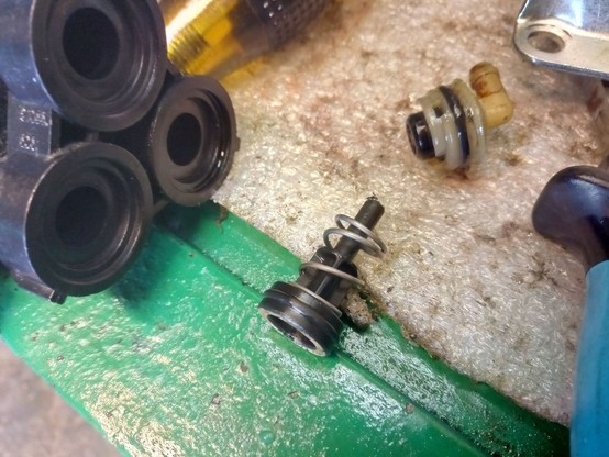 Black plastic triple-head pump housing on the left, cap with seal and detergent injector at the top, pressure pin with return sprink in the center. The pin is visibly frayed and worn.