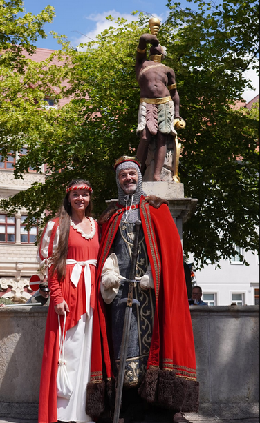 Foto vom Eisenberger Markt: Ein mittelalterlich verkleidetes Paar steht lächelnd vor dem M*Brunnen mit der kolonialistischen Statue