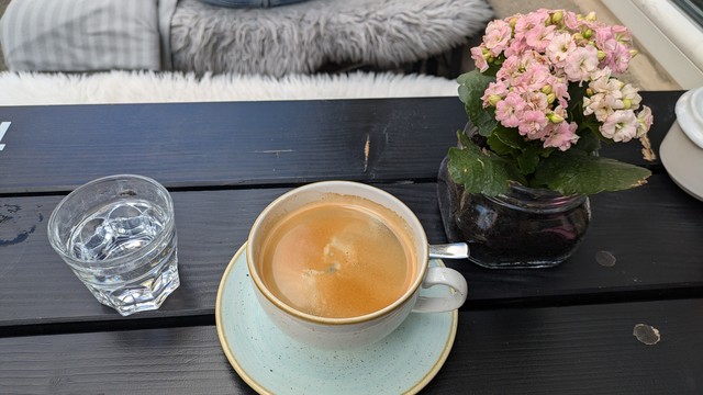 Große Tasse schwarzer Kaffee, kleines Glas Wasser auf einem Cafétisch neben einem pinkfarbenen Blümchen