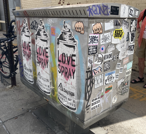 On a sidewalk, a metal bin is covered with stickers and graffiti. Among them are 3 copies of a (stenciled and sprayed?) picture of an aerosol can of LOVE SPRAY/CAUTION: CONTENTS MAY BE CONTAGIOUS 