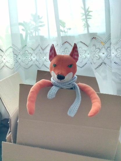 A fox in a box. The fox is a selfmade stuffed plushie in orange with a white neck, and big black nose. He's wearing a crocheted blue scarf and has green eyes. His long arms are hanging over the edge of the open cardboard box lid.