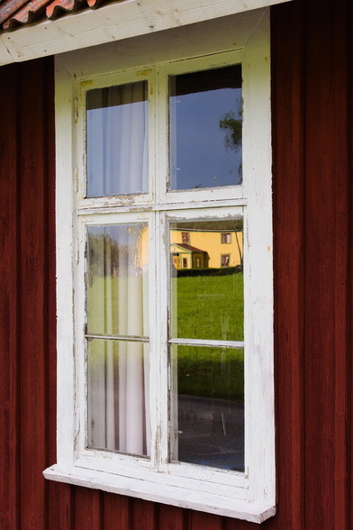Ein altes weisses Fenster in einem typisch rot gestrichenden Holzhaus. Über dem Fenster beginnt angeschnitten das Dach, weil das Bild schräg aufgenommen wurde, damit sich das gelbe Haus gegenüber in den Scheiben spiegelt.
Das gelbe Haus und die grüne Wiese davor wird von der Morgensonne angestrahlt. Das Fenster im roten Haus liegt dementsprechend im Schatten.