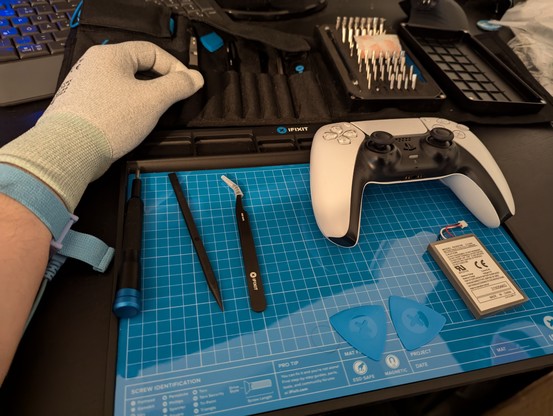 A white PS5 controller is placed on a reflection blue plastic mat with several libes on it.
Next to it is a small controller battery, screwdriver, plastic picks, tweezers and a small, lengthy plastic stick.

On top a small kit of bits and other accessory is visible.

My hand on the left is visible wearing bright anti-static gloves and an anti-static wristband.

Seberal visible pieces where the blue iFixit logo resembling the top of a Phillip's screw slightly.