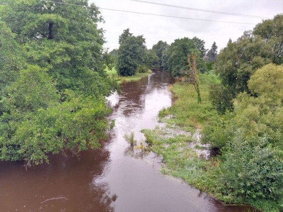 A normally small river that is swollen by rain. Murky brown waters rush downstream, the water level is very high and has flooded some of the shallow embankment.