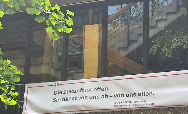 Ein Banner an einem Plattenbau wird von Sonnenstrahlen angestrahlt. Auf ihm steht "Die Zukunft ist offen. Sie hängt von uns ab - von uns allen." - Karl Popper (1902 bis 1994) Wissenschafts und Sozialphilosoph