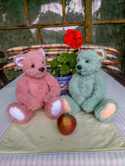 Pink bear with white ears and footpads, and minty green bear with pastel orange ears and footpads. Sitting on a table, apple in front of them, nice red flower behind. Looking cute.