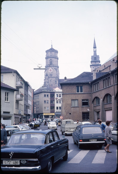 Bild aus den 1960iger. Blick vorbei an der Markthalle in Stuttgart auf die Stiftskirche. Vor der Markthalle stehen und fahren Autos. Heute ist dort eine Fußgängerzone und Aufenthaltsqualität.