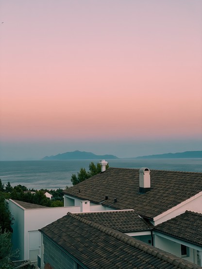 Blick über ein paar Dächer mediterraner Häuser aufs Meer mit entfernten Inseln und rosa gefärbtem Himmel 