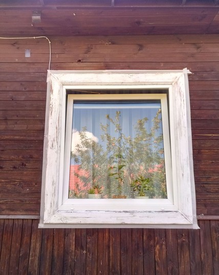 White PVC window set in a log house with brown exterior planking. The siding was also white, but flaking paint has been scraped and sanded, some caulking applied in the mitered corners and on the bottom joint between window and siding. The wood looks old and manky. Masking tape surrounds the outer edges.