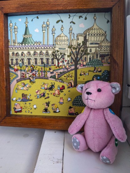 A small pink bear with grey and white dotted ears and paws, sitting in front of a wooden picture frame that contains a postcard print with a drawing of Brighton pavillion and gardens with lots of characters frolicking on the grass. He's got traditional button joints in various colours and a hand stitched mouth and nose. The eyes are small black pins.