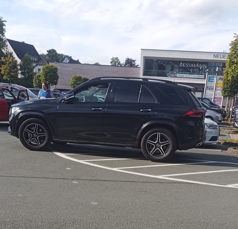 Dicker schwarzer SUV steht halb auf dem Parkplatz, halb auf dem gestrichelten und mit dem Reifen halb in der Fahrspur auf einem Supermarktparkplatz.