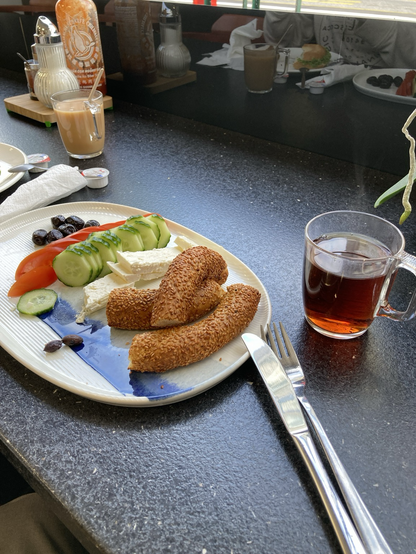 Beside a glass of tea on a counter, a plate with a quartered sesame-seed-encrusted ring bread, white cheese, sliced cucumber and tomato, oil-cured olives 