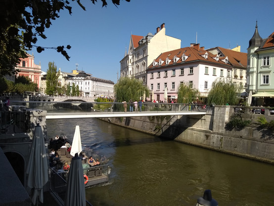 Ljubljana: Ausblick auf den Fluß Ljubljanica in der Altstadt. Im Wasser ein Ponton auf dem Leute sitzen. Im der Bildmitte eine neue Fußgängerbrücke mit durchsichtigen Glasgeländer. Im Hintergrund die Brücken des zentralen Altstadtplatzes. Der Fluß selbst ist hier durch Mauern eingegrenzt. Oberhalb davon auf beiden Seiten Promenaden. Alles autofrei seit 2007. Links und rechts des Flußes alte Häuser und Kirchen aus der österreichischen K.U.K. Zeit