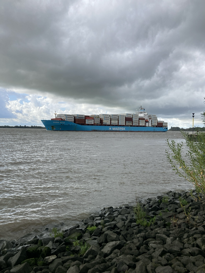 Under a dark gray sky with some blue on the horizon, the Maersk Baltur, a blue container ship loaded six containers high