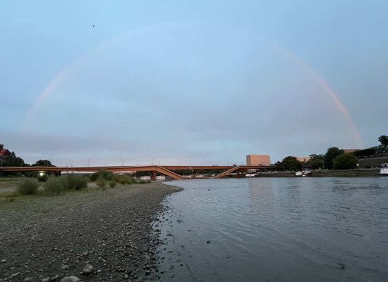 Bild vom Flussufer in Fluissrichtung, links Steinufer/-strand und Gebüsche. Weit entfernt im Hintergrund eingestürzte Brücke sowie Regenbogen im Himmel darüber