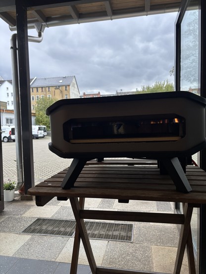 A pizza oven on a wooden stand, positioned in an indoor area with large glass windows. Outside, there are cloudy skies and a view of a street with buildings and parked vehicles.