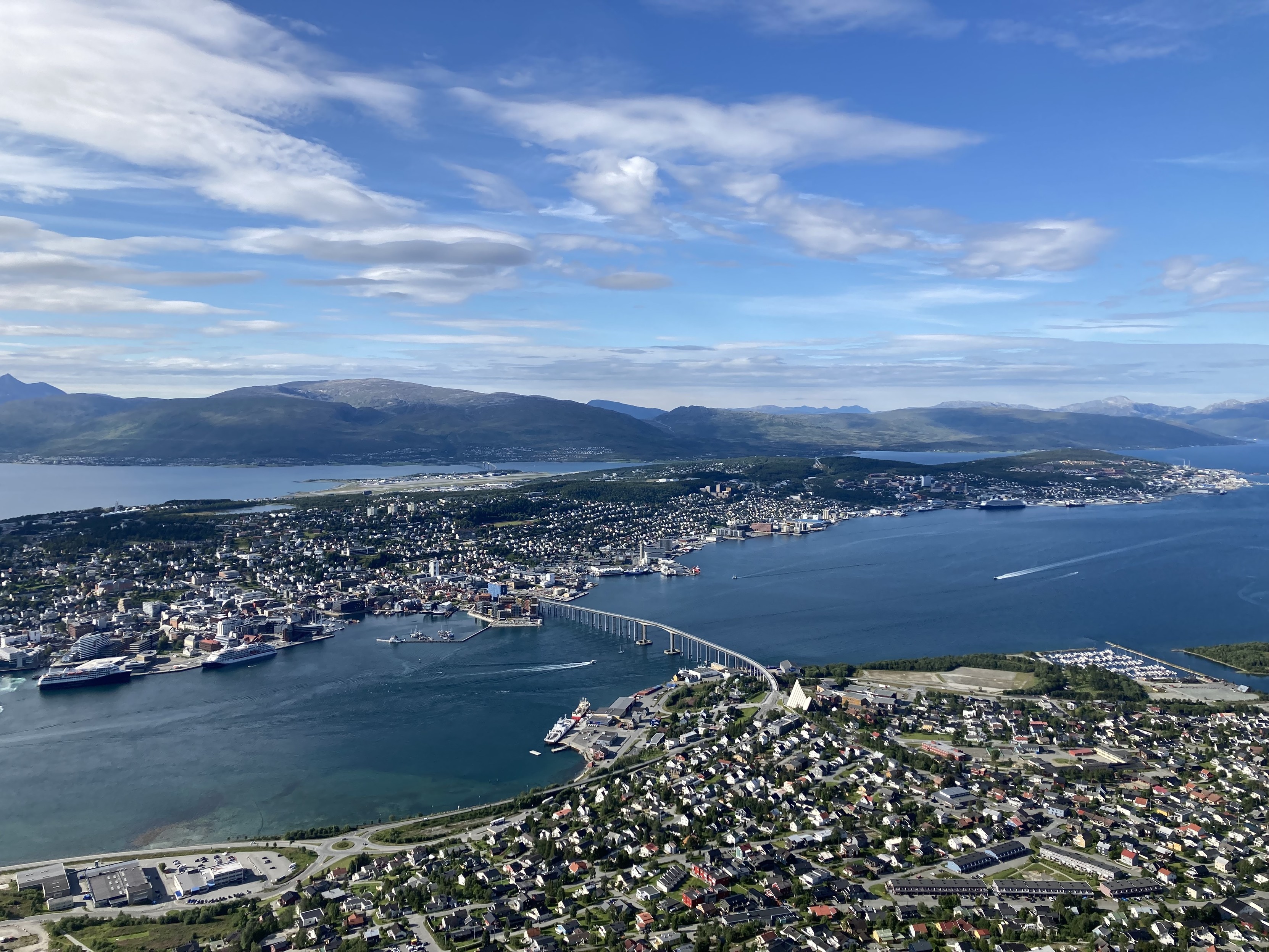 Das Bild zeigt die Stadt Tromsø von einem Berg aus fotografiert. Zu sehen sind die beiden bewohnten Landteile, die von Wasser getrennt und mit einer Brücke verbunden sind. Im Hintergrund ist eine Bergkette zu sehen. Der Himmel ist blau, mit einzelnen hellen Wolken. Das Bild zeigt die Stadt Tromsø von einem Berg aus fotografiert. Zu sehen sind die beiden bewohnten Landteile, die von Wasser getrennt und mit einer Brücke verbunden sind. Im Hintergrund ist eine Bergkette zu sehen. Der Himmel ist blau, mit einzelnen hellen Wolken.
