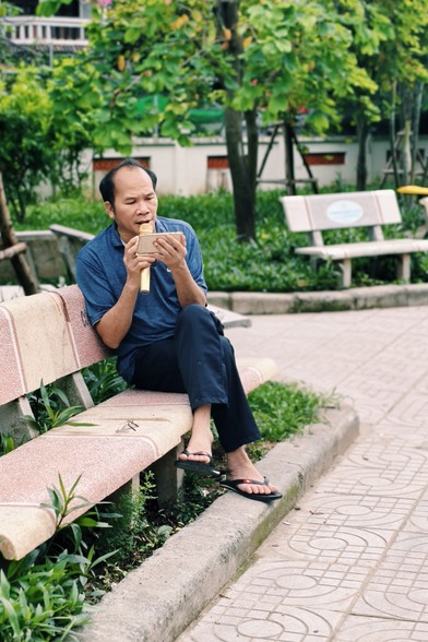 On a solo cement park bench, green trees around, a men sings Karaoke in a golden mic connected to a phone 