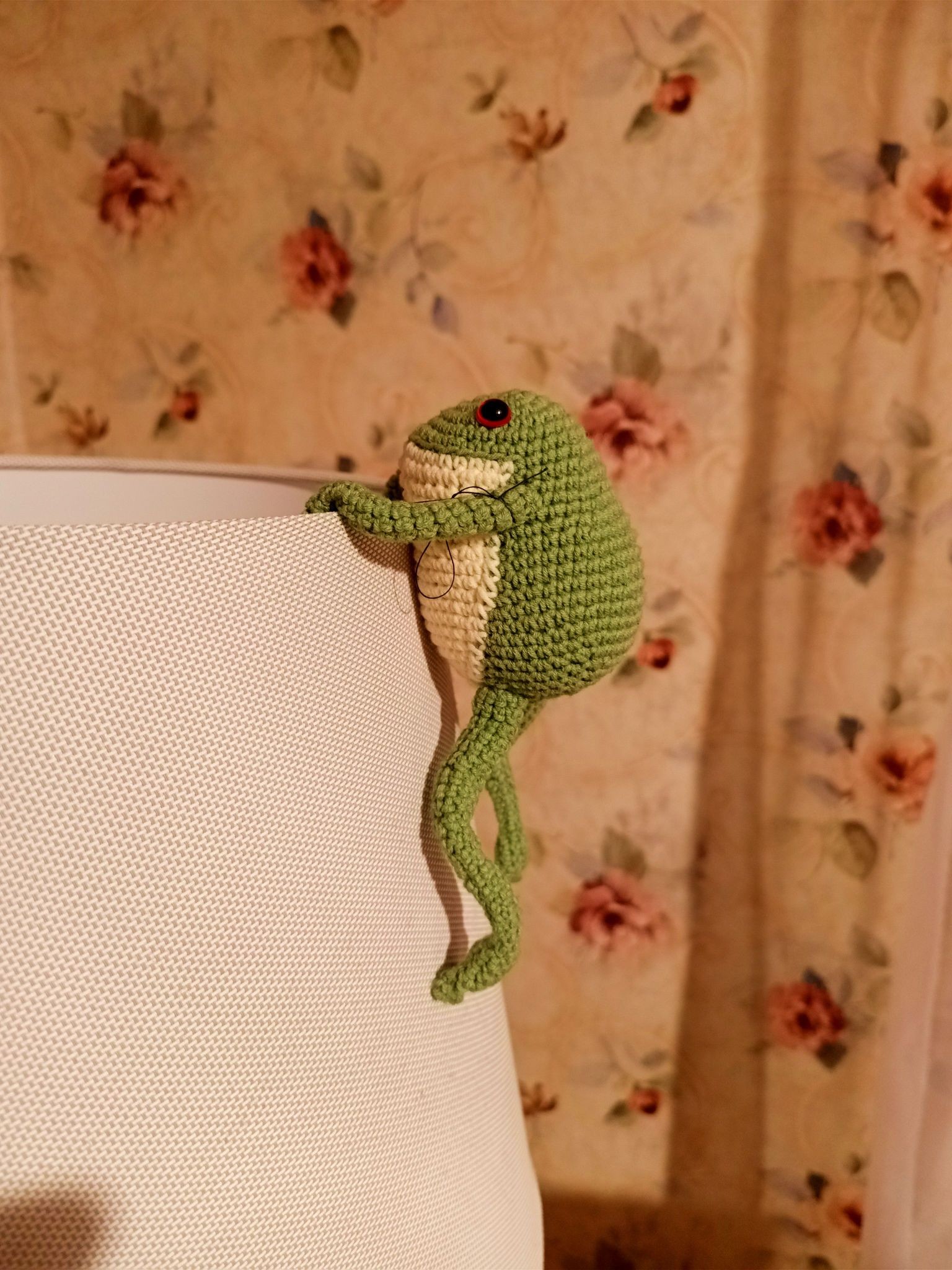 A crochet frog from green and white yarn with glossy eyes and log limbs. It's climbing up the outside of a lampshade, hands holding on to the upper rim.