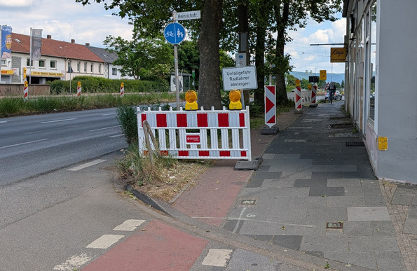 Ein schlechter, schmaler Radweg entlang einer zweispurigen Stadtstraße ist mit eine rot-weißen Gatter versperrt. Neben dem Zeichen 241, welches eine Benutzungspflicht ausweist, steht ein Schild "Unfallgefahr Radfahrer absteigen".