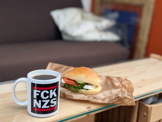 Eine Tasse Kaffee. Auf der ansonsten weißen Tasse steht in weißer Schrift auf schwarzem Grund FCKNZS. Neben der Tasse liegt auf einer Papiertüte ein Käse-Tomate-Brötchen 