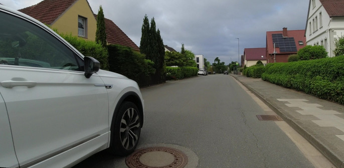 Einzelbild einer Dashcamaufnahme: ein weißer SUV überholt einen Fahrradfahrer. Das rechte Vorderrad ist mit der Außenkante an der äußeren Kante eines Gullideckels.