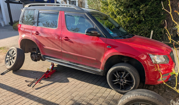 Ein roter Skoda Yeti - Ausstattungsvariante "Monte Carlo" in der Farbvariante schwarz-rot - steht mit der rechten Hinterseite per Wagenheber hochgebockt auf einer gepflasterten Fläche. Der rechte Hinterreifen ist demontiert und lehnt am hinteren, rechten Kotflügel.
