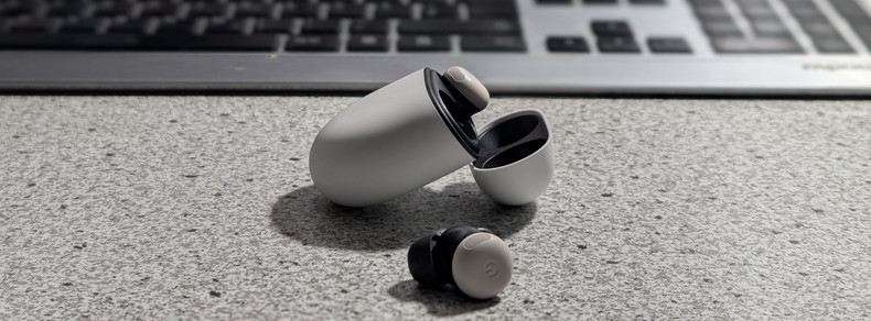 Google Pixel Buds 2 Man sieht im Vordergrund eine weiße, rundliche Schatulle zur Aufbewahrung von zwei In-Ear-Kopfhöreren. Einer der kleinenStöpsel ist an seinem Platz in der Schatulle, ein Hörer liegt vor der Hülle auf einer gesprenkelten Tischplatte. Im Hintergrund ist verschwommen ein Teil einer Tastatur zu sehen.