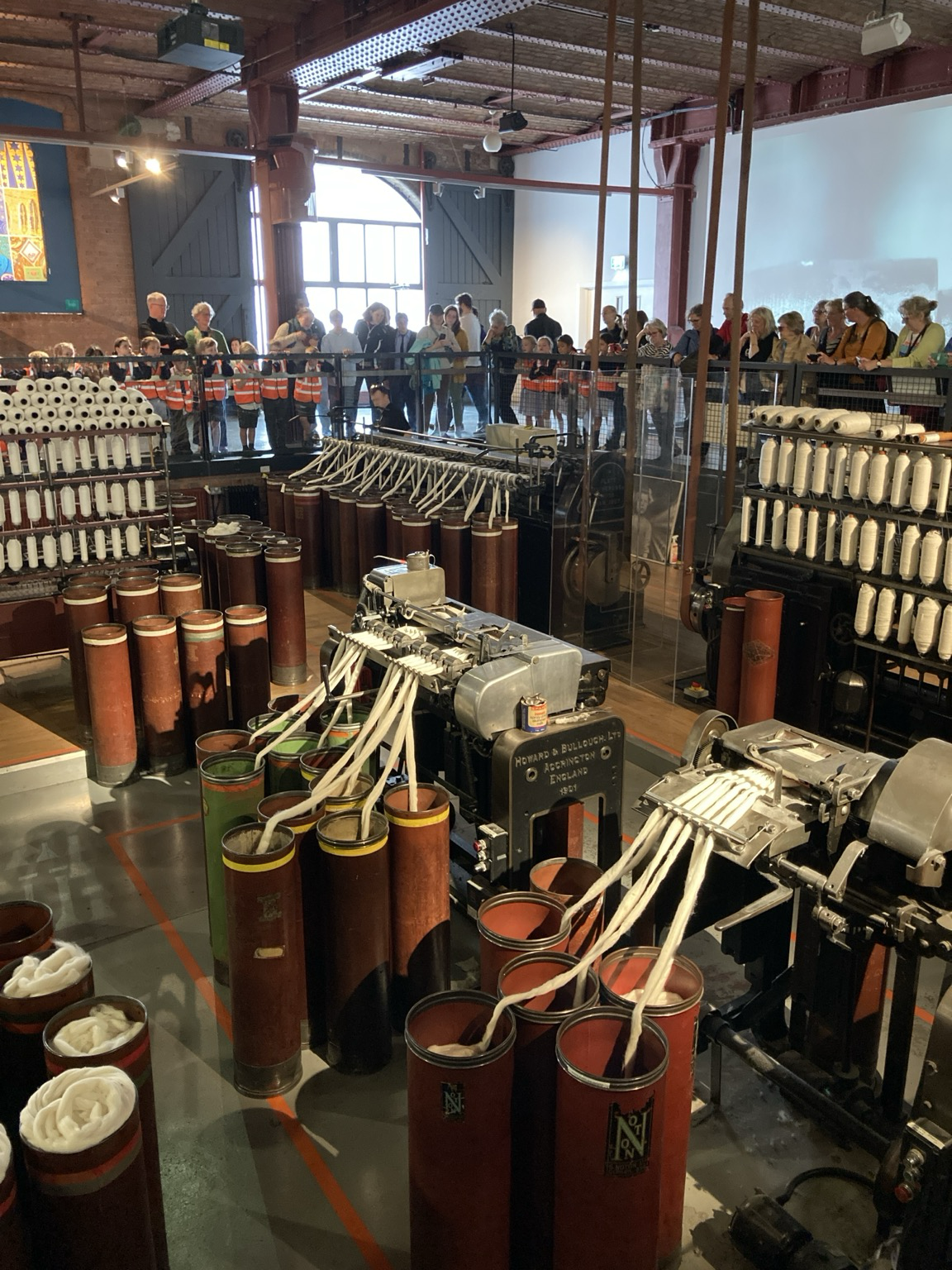 In the pit, spinning machines and machines that remove impurities from thick skeins of cotton; around wrim of the pit behind railings people look on.
