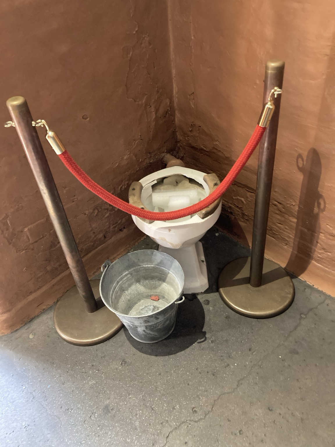In a corner of a cell, a commode filled apparently with styrofoam blocks and set behind a velvet rope