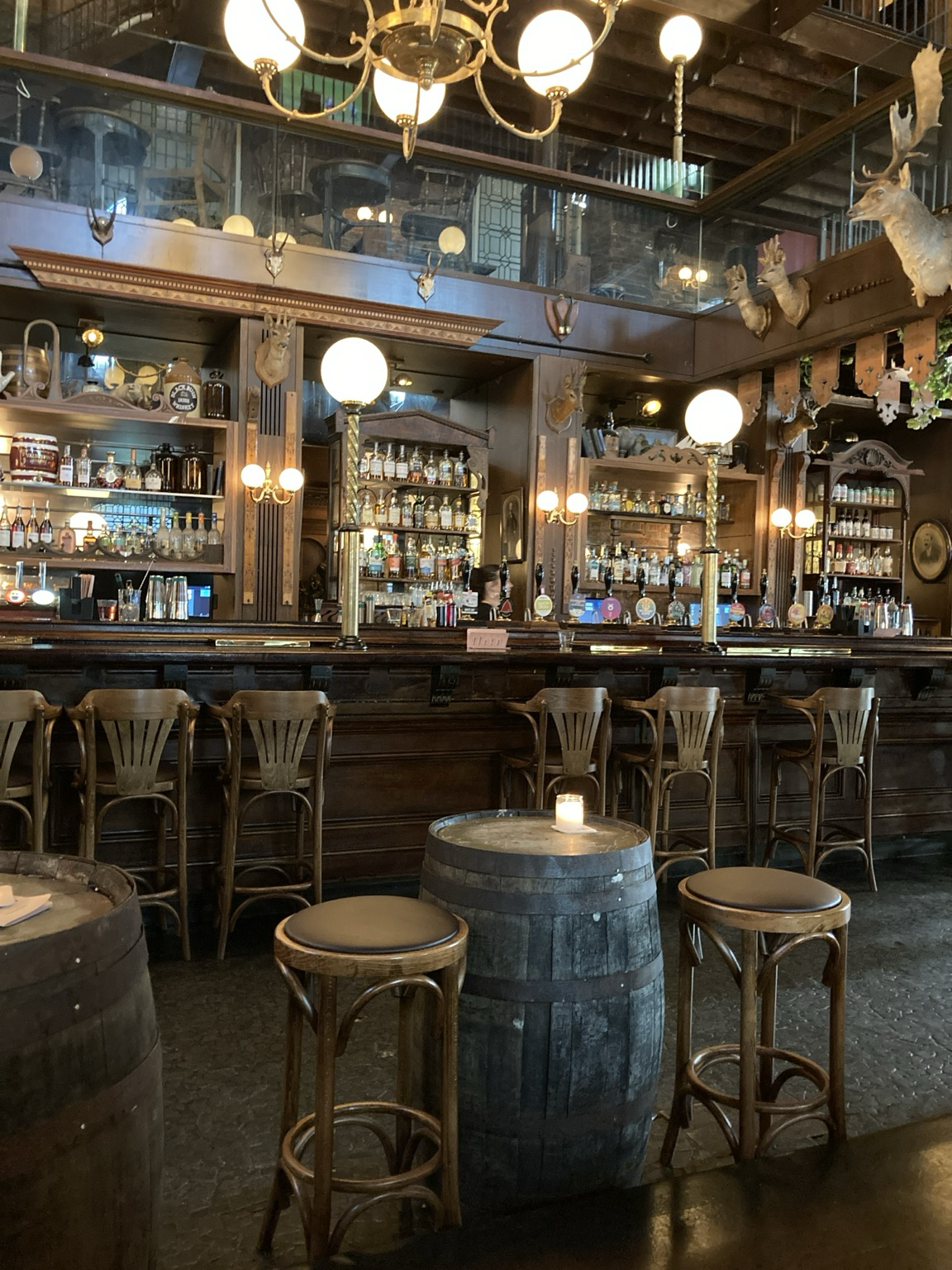 Looking toward the long bar past barrel tables and stools. There are mounted deer heads.