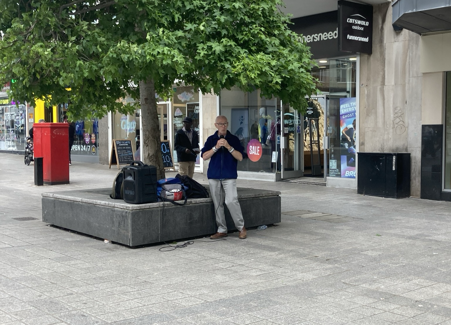 Elderly guy in polo shirt clutching a mike under a tree in a shopping plaza