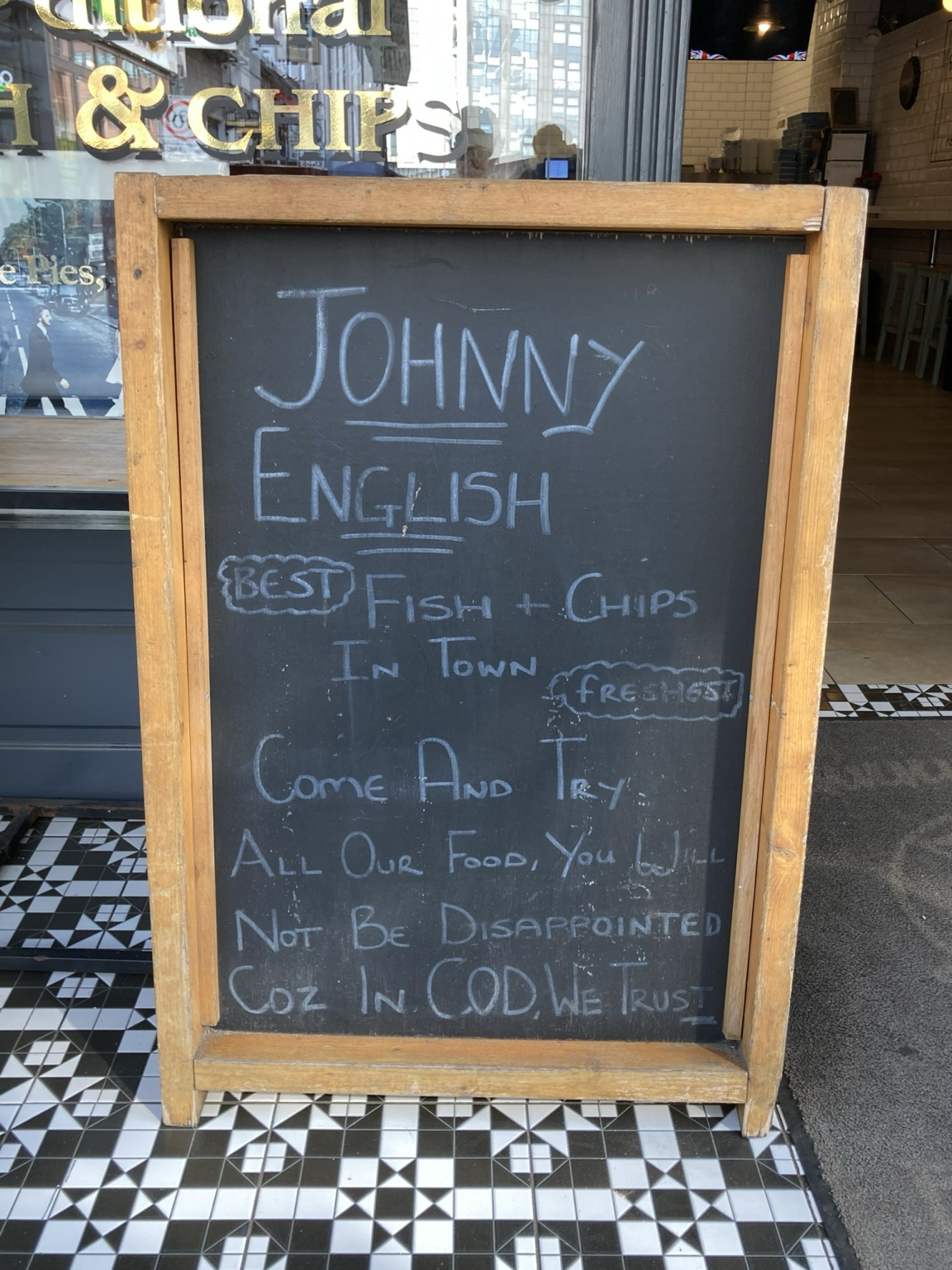 On a blackboard: JOHNNY ENGLISH BEST FISH + CHIPS In TOWN FRESHEST COmE AND TRY. ALL OuR FooD, You Will NOT BE DISARFOINTED Coz IN. COD .WE TRUST