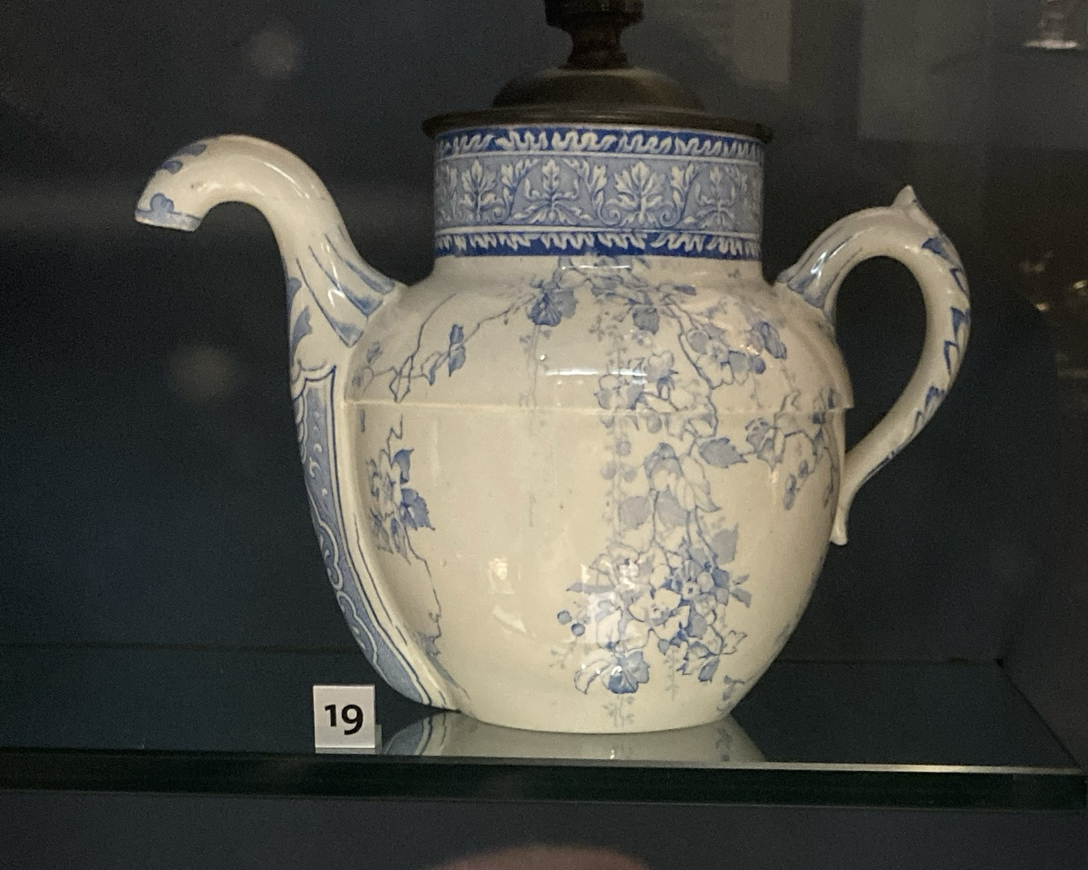 White teapot with downward pointed spout, blue trace work floral design, and a metal lid and knob