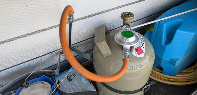 Das Innere eines Deichselkasten im Wohnwagen. Man sieht in der Mitte eine graue Gasflasche mit einem Druckminderer mit grünem Etikett. Rechts ist die Hälfte eines mobilen, blauen Grauwassertanks zu erkennen.